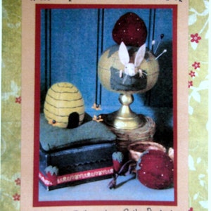 May include: A vintage-style pincushion pattern featuring a white rabbit peeking out of a red strawberry pincushion, a beehive pincushion, and a small wooden box. The pattern is titled "Pinkee Pos From The Berry Patch" and is by Cathy Pendleton, The Cheswick Company.