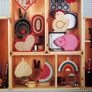 Puede incluir: Una vitrina de madera llena de varios salvamanteles de ganchillo en diferentes formas y colores. Los diseños incluyen un corazón, una manzana, una rodaja de sandía y un arcoíris. El texto "Crocheted Potholders" es visible en la parte superior.