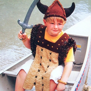 May include: A young boy dressed as a viking, wearing a brown knit hat with horns, a brown and tan knit vest, and white shorts. He is holding a silver toy sword and sitting in a canoe.