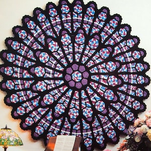 Puede incluir: Un afgano circular y galardonado con un diseño de vidriera. La pieza de ganchillo presenta un borde negro, con secciones moradas, azules y rojas. El texto "Award Winning Afghans" está en la parte superior.