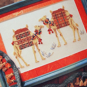 Puede incluir: Arte de punto de cruz de dos camellos con alforjas decorativas y borlas coloridas, enmarcado en un marco de madera azul. La obra de arte está sobre un fondo blanco con acentos rojos y naranjas. Un adorno decorativo con borlas está en primer plano.