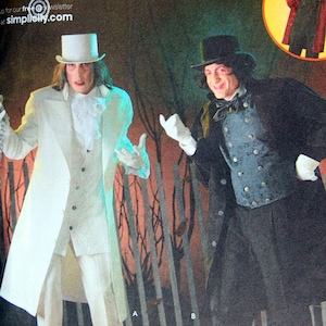 Op de afbeelding: Twee mannen in kostuums voor een Halloween-feest. De man links draagt een wit pak met een lange jas en een hoge hoed. De man rechts draagt een zwart pak met een lange jas en een hoge hoed. De achtergrond is een donker bos met bomen en takken.