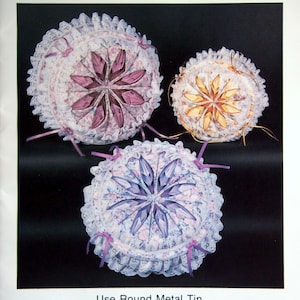 May include: Three round, white, lace-trimmed fabric cushions with a daisy-like design in shades of purple, yellow, and blue. The text "DAISY DECORATOR TIN" is at the top of the image. The text "NO SEWING REQUIRED" is below the cushions. The text "Use Round Metal Tin." is below the text "NO SEWING REQUIRED". The text "Fast - Easy - Fun." is below the text "Use Round Metal Tin."