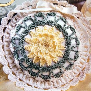 May include: A crocheted floral sachet with a white lace border and a yellow and green flower design. The sachet is tied with a white ribbon and has the text "Sachet Blossom" above it.