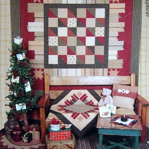 Peut inclure: Un livre intitulé "Simple Gifts" par Kansas Troubles Quilters, présentant une courtepointe avec un motif patchwork rouge, blanc et marron. La courtepointe est exposée sur un canapé en bois avec un ours en peluche et une petite table avec une bougie.