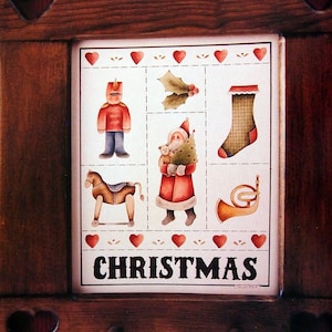 May include: A wooden frame with a heart-shaped cutout on each side. The frame contains a white tile with a Christmas-themed illustration and the word "CHRISTMAS" printed in black. The tile is divided into nine squares, each containing a different Christmas image, including a toy soldier, a stocking, a Santa Claus, and a rocking horse.