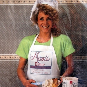 Puede incluir: Una mujer con un gorro de cocinero blanco y una camisa verde con un delantal blanco que dice "Mom's Diner Serving 24 hours a day" sostiene una espátula y un plato de pastel con una bola de helado y una rebanada de pastel. Hay un pastel en un plato y un recipiente de helado en una mesa de madera.