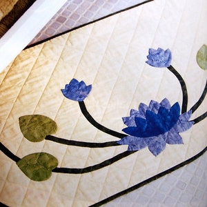 May include: Close-up of a quilted fabric with a floral design. The fabric is a light beige color with a diamond pattern. Dark blue lotus flowers and green leaves are appliqued onto the fabric, with black stems. The design is likely for a table runner or similar home decor item.