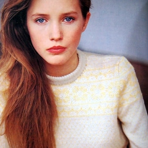 May include: A young woman with long, straight, auburn hair is wearing a white sweater with a yellow and white pattern.