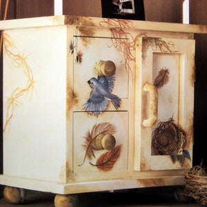 May include: A white painted wooden cabinet with a bird and feather design. The cabinet has three drawers and a door with a handle. The cabinet is on wheels and has a distressed finish.