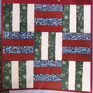 Puede incluir: Un patrón de bloque de colcha con el título "JULY Roman Square". El bloque está formado por nueve cuadrados más pequeños, cada uno con un patrón diferente de tela roja, blanca, azul y verde.