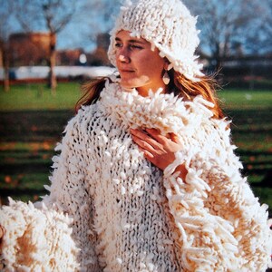 May include: A white knitted poncho with a fluffy texture and fringe detail. The poncho is worn with a white knitted hat.