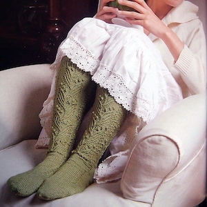 Puede incluir: Una mujer sentada en un sillón blanco, vestida con un vestido blanco con ribete de encaje y calcetines hasta la rodilla de color verde oliva. Los calcetines presentan un patrón de punto detallado. Sostiene una taza verde.