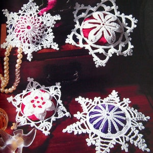 May include: Four white crocheted snowflake sachets with different colored fabric centers. The centers are red, purple, and two different shades of pink. The snowflakes are displayed on a wooden surface with a pearl necklace.