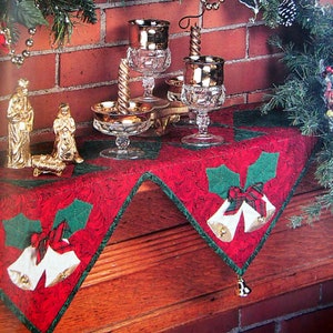 May include: A festive Christmas display featuring a red and green table runner with bell and holly designs. Crystal goblets with gold rims and candles are arranged on the runner. A garland with poinsettias and ornaments adorns the brick background.