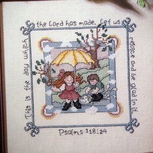 Könnte beinhalten: Kreuzstich-Stickerei in einem braunen Holzrahmen. Das Design zeigt zwei Kinder unter einem Schirm mit gelber Kuppel. Der Text lautet "This is the day which the Lord has made. Let us rejoice and be glad in it. Psalms 118:24"