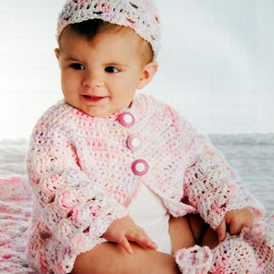 以下が含まれることがあります： ピンクと白の編み物カーディガンを着て、白いボタンとそれに合わせた編み物の帽子をかぶった赤ちゃん。赤ちゃんはピンクと白の編み物の毛布の上に座っています。
