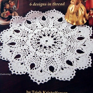 May include: A white, crocheted doily with a pineapple design, displayed on a dark surface. The image is from a crochet book titled "Heirloom Pineapple Doilies" by Trish Kristoffersen. The book cover features decorative figurines.