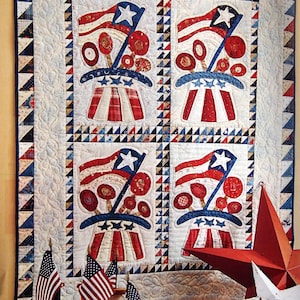 Peut inclure: Une courtepointe patriotique avec quatre blocs rouge, blanc et bleu représentant un drapeau et un chapeau avec des étoiles. La courtepointe est encadrée et accrochée à un mur. Il y a trois bobines de fil et trois drapeaux américains devant la courtepointe.