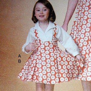 May include: A young girl wearing a white collared shirt and a red and white floral print jumper dress. The dress has a pleated skirt and suspender straps. The girl is smiling and wearing white socks and shoes. A matching dress is partially visible.