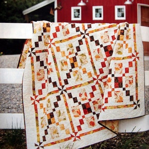 Op de afbeelding: Een patchwork quilt met een herhalend patroon van crème, oranje, rode en bruine vierkanten. De stof toont een printmotief van herfstbladeren en geometrische vormen, met kleine windmolenaccenten op de hoeken van de bloksecties.