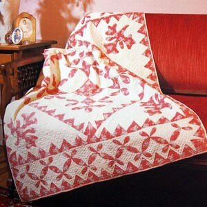 Könnte beinhalten: Eine weiße und rosafarbene Steppdecke mit Blumenmuster liegt über einem roten Sofa. Die Steppdecke hat einen gewellten Rand und ist aus Baumwollstoff gefertigt.