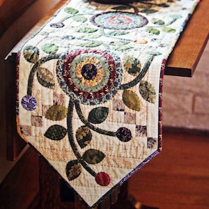 Op de afbeelding: Een witte tafelrunner met een bloemenontwerp met groene bladeren, bruine ranken en kleurrijke cirkels. De runner ligt op een houten tafel met twee houten kommen erop.