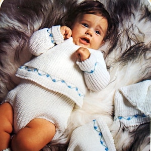 May include: A white knitted baby outfit with blue embroidered flowers. The outfit includes a sweater, trousers, and a bonnet. The outfit is laid out on a white furry surface. No. 5128-187 (ITO-KNIT)