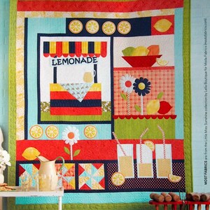 Op de afbeelding: Een kleurrijk quilt met een limonadestand, citroenen, bloemen en glazen limonade. De quilt is gemaakt met verschillende stoffen en patronen. De quilt hangt aan een muur met een witte bakstenen achtergrond. De quilt is omlijst door een groene rand.