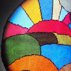 May include: A round rug with a colourful geometric design. The rug is made of a soft, plush material and features shades of blue, green, orange, yellow, pink, and brown.