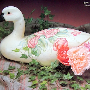 May include: A white wooden duck painted with pink and green flowers. The duck is lying on a fabric surface with green ivy.
