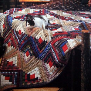 Op de afbeelding: Een patchwork quilt met een geometrisch patroon in rode, blauwe en beige tinten bedekt een bed. Een zwart-witte kat ligt op de quilt. De slaapkamer heeft een rustieke, hutachtige uitstraling, met houten meubels en een warme sfeer.