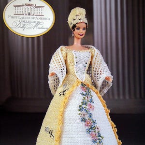 May include: A doll from the First Ladies of America Collection, Dolly Madison, in a crocheted gown. The white dress has yellow trim and floral embroidery. The doll also wears a matching bonnet and shawl.