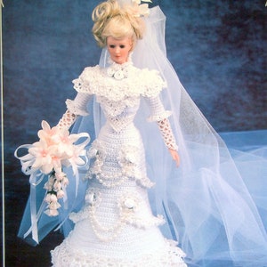 Puede incluir: Muñeca vestida con un vestido de novia blanco de ganchillo hecho a mano, con velo y ramo. El vestido presenta un corpiño detallado, mangas largas y una falda escalonada. La muñeca sostiene un ramo de flores rosas y tiene el pelo rubio recogido.