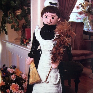 Könnte beinhalten: Eine gehäkelte Puppe, die als Dienstmädchen verkleidet ist, trägt eine weiße Schürze und ein schwarzes Kleid und hält einen Besen und einen Staubwedel. Die Puppe steht vor einem Stuhl und einem Blumenarrangement.