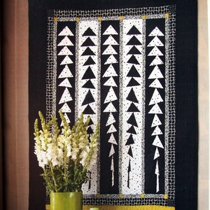 May include: A black and white quilt with a geometric pattern of triangles and dots. The quilt is framed and hanging on a wall. A vase of white flowers sits on a black table in front of the quilt. The text "Turn to page 16 for more designer Kay Capps Cross" is visible at the bottom of the image.