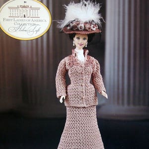 May include: A doll from the First Ladies of America Collection, dressed in a crocheted pink suit and skirt, a white blouse, and a hat with feathers. The doll has a detailed face and is posed against a blurred background.