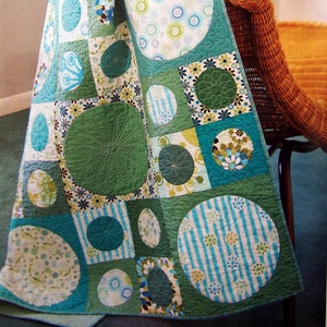 May include: A green and blue patchwork quilt with a circular pattern. The quilt is made of different fabrics with various patterns, including circles, stripes, and flowers. The quilt is draped over a wicker chair.