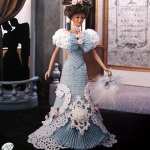 May include: A doll in a light blue crocheted gown with white lace trim, a matching fan, and a feathered headdress. The doll has a pearl necklace and is posed in a room with a decorative door and a calendar for June.