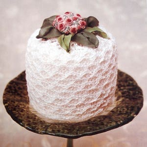 May include: A white crocheted toilet paper cover with a pink flower embellishment on top. The cover is sitting on a green pedestal.