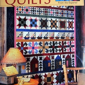 Puede incluir: Una portada de libro titulada "Basic Guide to Rotary Cut Quilts" que presenta un edredón colorido con varios patrones geométricos, incluyendo estrellas, casas y triángulos. El edredón tiene un borde rosa y se muestra sobre un fondo de madera.