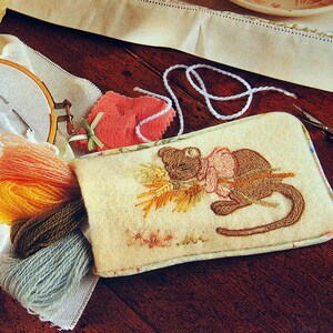 May include: A beige needlepoint project bag with an embroidered image of a brown cat holding a bundle of wheat. The bag is surrounded by embroidery supplies, including a hoop, yarn, and scissors.