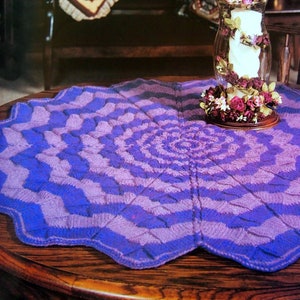 May include: A purple and blue crocheted table runner with a circular design. The runner is on a wooden table with a vase of dried flowers in the center.