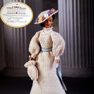 May include: A doll from the First Ladies of America Collection, Edith Roosevelt, in a cream crocheted dress, hat, and purse. The hat has colourful floral accents. A blue ribbon belt and sash detail the dress.