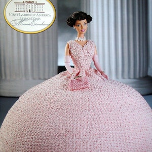 May include: A Barbie doll from the First Ladies of America Collection, Mamie Eisenhower edition, in a light pink crocheted gown. The doll has dark hair and wears a pearl necklace, earrings, and gloves. The dress features a full skirt and a fitted bodice.