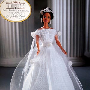May include: A doll from the First Ladies of America Collection, dressed in a white lace gown with a tulle veil. The doll has dark hair, a tiara, and pearl jewellery. The doll is positioned in front of a blurred background.