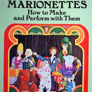 May include: A vintage book cover with the title "Marionettes: How to Make and Perform with Them" by Leonard Suib & Muriel Broadman. The cover features a colorful illustration of a puppet show with various puppets, including a clown, a magician, and a lady in a green dress.