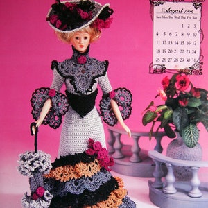 May include: A doll in a crocheted Victorian-style dress, hat, and parasol. The dress features layers of black, orange, and grey crochet. The hat and dress are adorned with pink accents. A calendar for August 1996 is in the background.