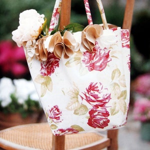 Puede incluir: Una bolsa de tela blanca con un estampado floral con rosas rojas y hojas verdes. La bolsa tiene rosetas de tela marrón alrededor de la parte superior y está sentada en una silla de madera.
