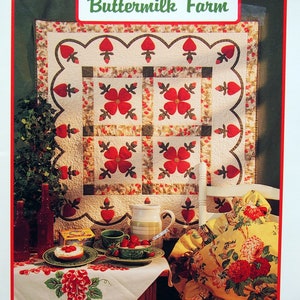 May include: A quilt with a floral design and the text "Buttermilk Farm" is displayed. The quilt features red flowers and heart-shaped accents. A table is set with a tablecloth, tea set, and a dessert. The image also includes a pillow with a floral pattern.